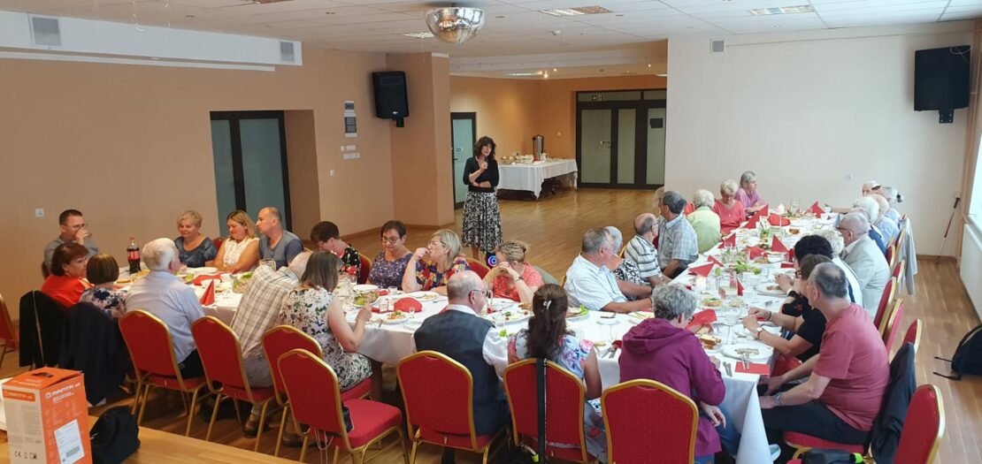 Widok na salę ze sceny. Przed sceną stoją stoły przy których siedzą uczestnicy spotkania. Przed stołami Pani Prezes stoi z mikrofonem